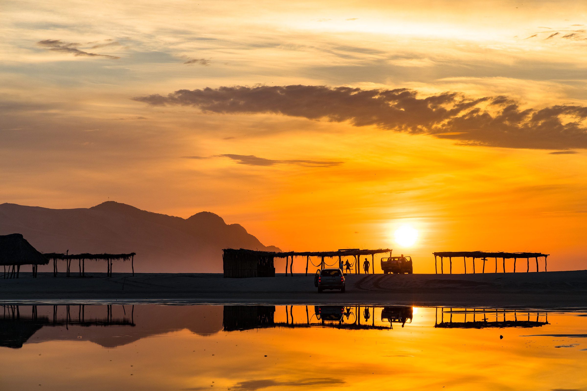 Sunset over the beach at Bamba, Salina Cruz — palapas and trucks silhouetted against the sun, mirrored in the wet sand