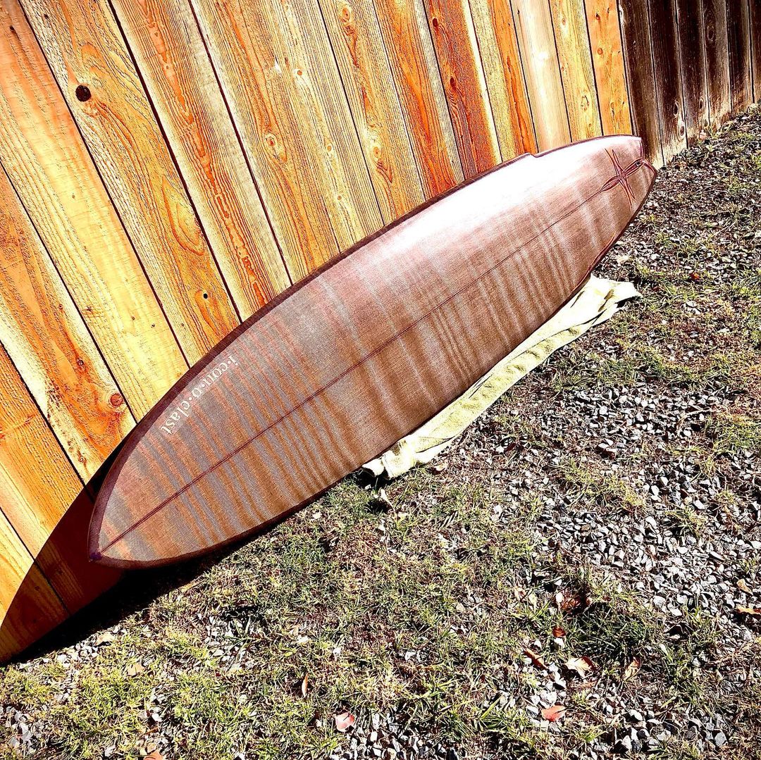 A wood-grained flax-basalt surfboard leaning against a sunlit fence