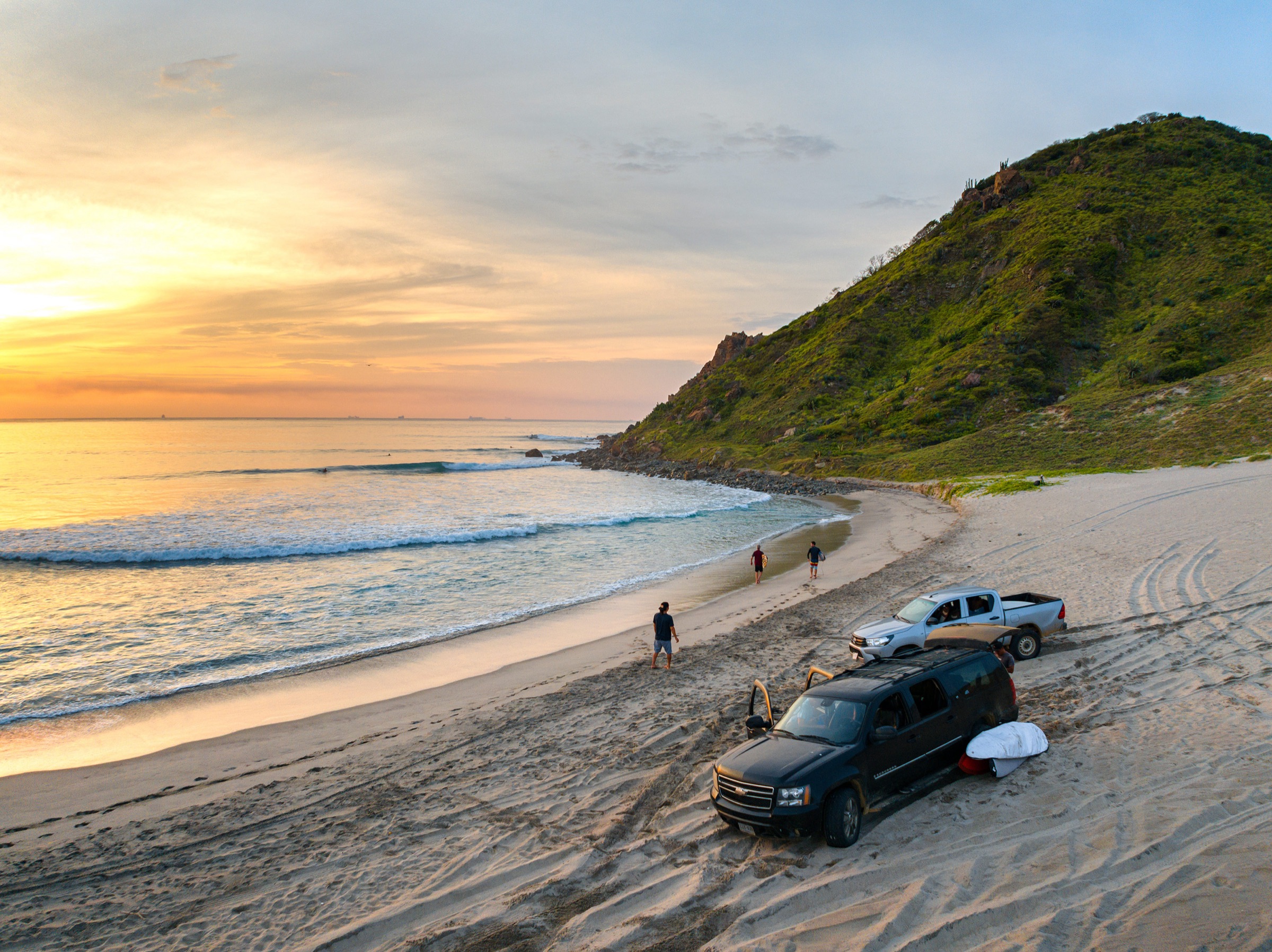 Golden-hour aerial of a beach at sunrise, surfers walking the shoreline
