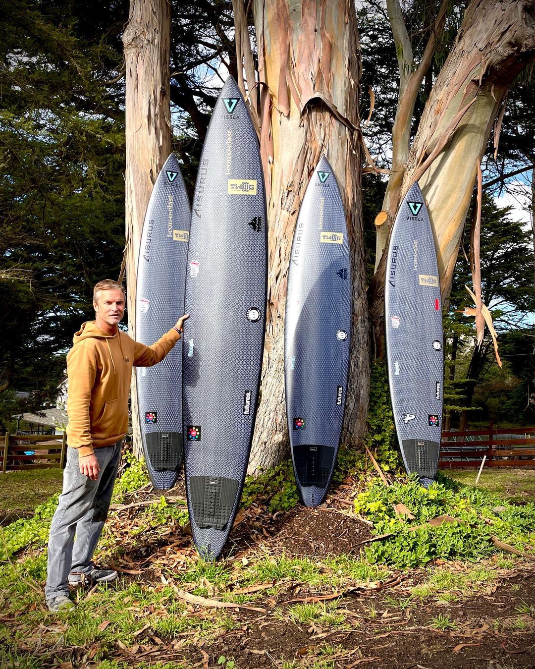 Mike Wallace standing beside four carbon big-wave guns against a eucalyptus tree